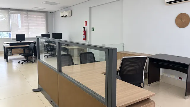Divisória de vidro e estações de trabalho, com relógio de parede e janelas ao fundo